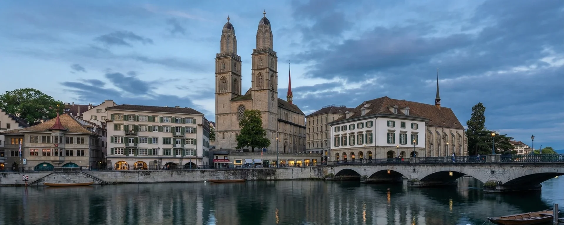 Großmünster in Zürich bei Abenddämmerung, umgeben von historischen Gebäuden und reflektiert im Wasser des Limmat-Flusses.